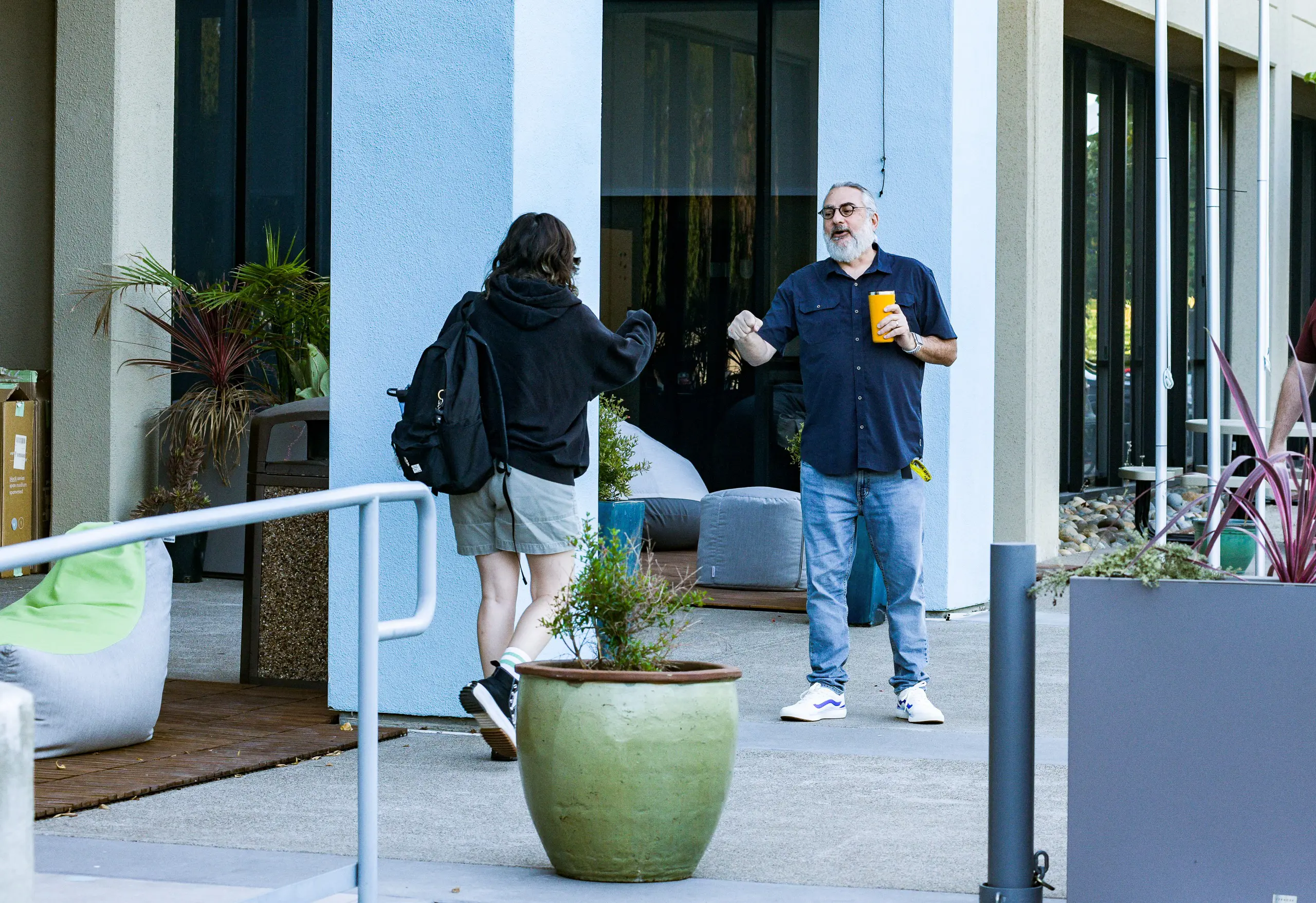Rabbi Dennis Eisner wearing a blue button-up shirt and blue jeans, holding an insulated mug in one hand, smiling at a student while holding out a fist bump. The back of a student in grey shorts and a 
