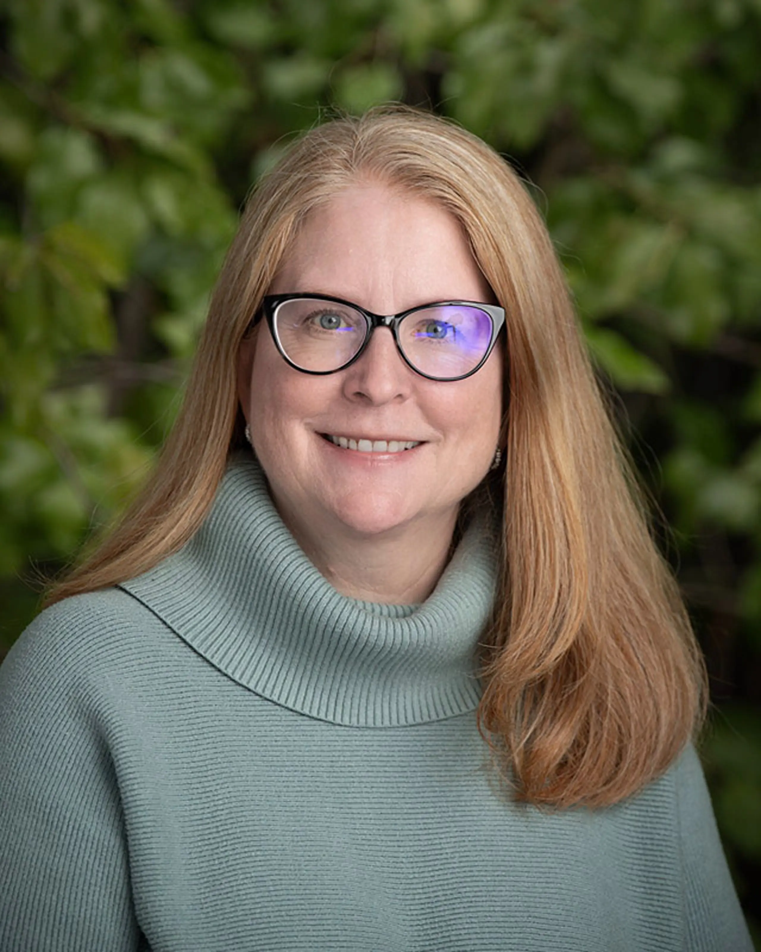 Portrait of Head of School Daisy Pellant wearing black glasses, a green and seafoam-colored sweater. She has mid-length ginger-orange straight hair and green eyes.