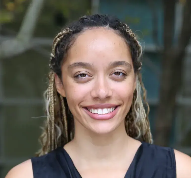 A smiling woman with braided hair stands in front of a tree, showcasing a cheerful expression and a natural backdrop.