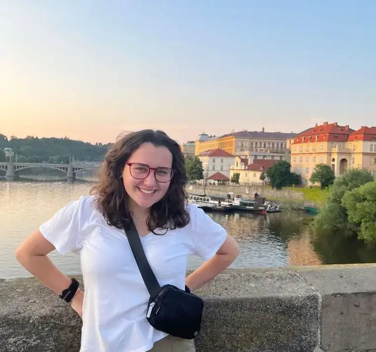 A woman poses on a bridge with a vibrant cityscape visible behind her, showcasing urban architecture and a bright sky.