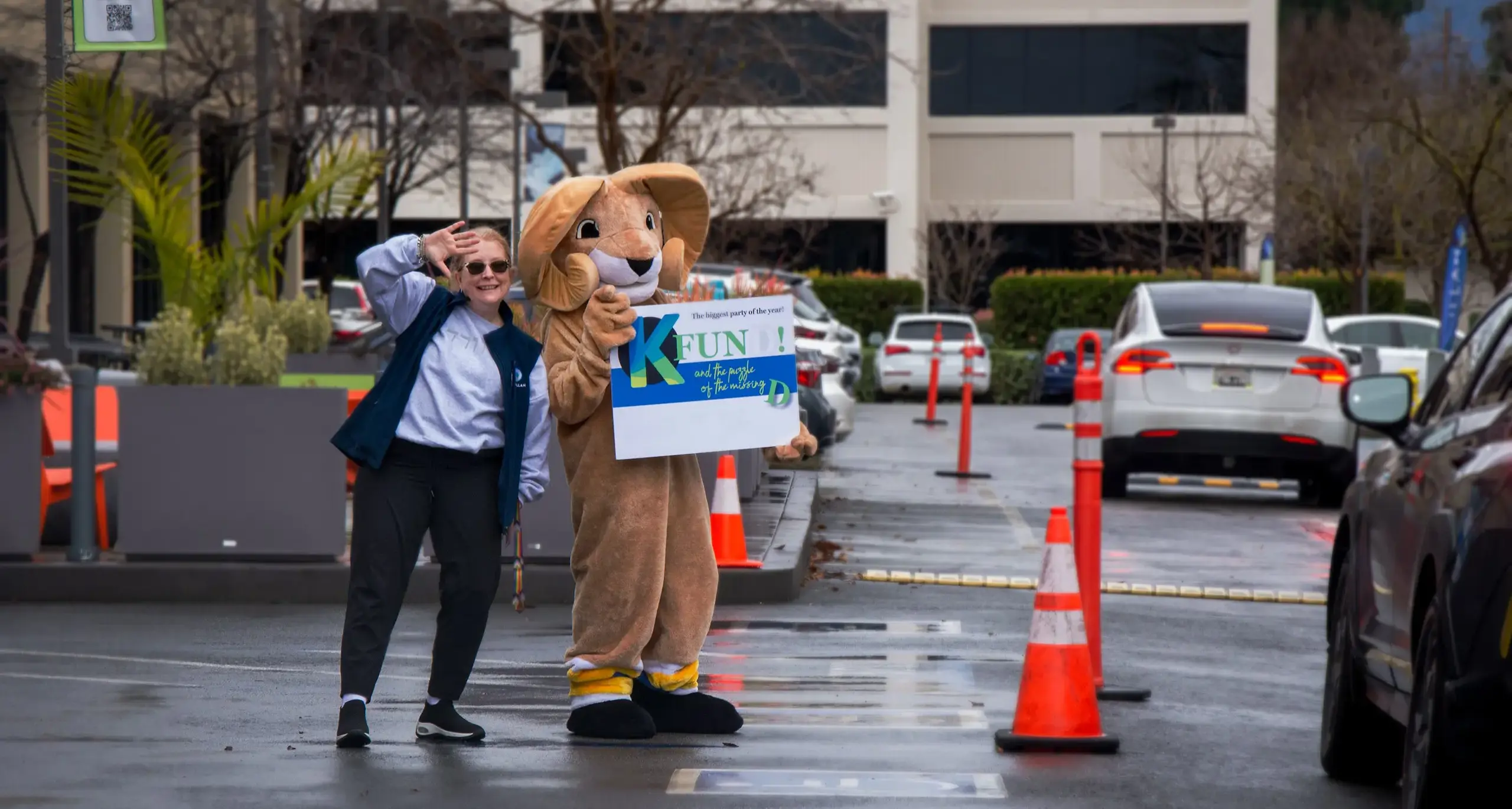 A volunteer in a blue jacket is smiling and waving alongside the mascot, a large character with a brown lion costume, both holding a sign that reads 'K-FUN!' in a parking lot with cars and orange traf