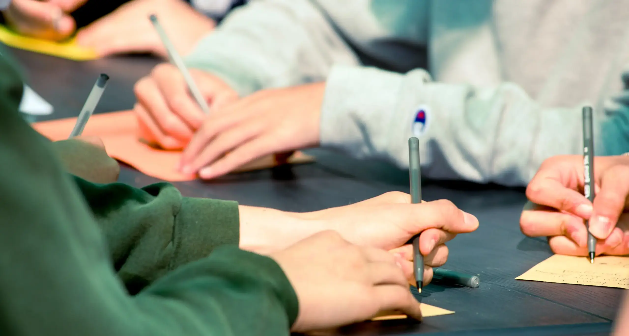 Three students are seen at a table in a classroom or study area, writing on pieces of paper. One student is wearing a green sweater, another student is wearing grey, and another student is cut out of 