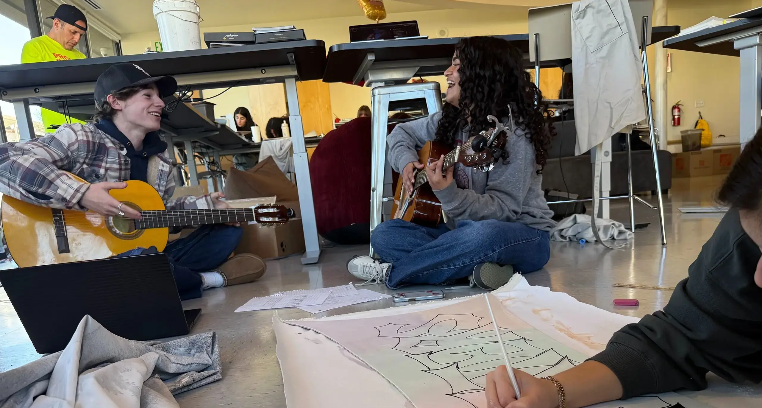 A group of students attentively sitting at tables in a classroom, engaged in learning activities. Two students are playing guitar. One student is painting and there is a teacher in the back painting.