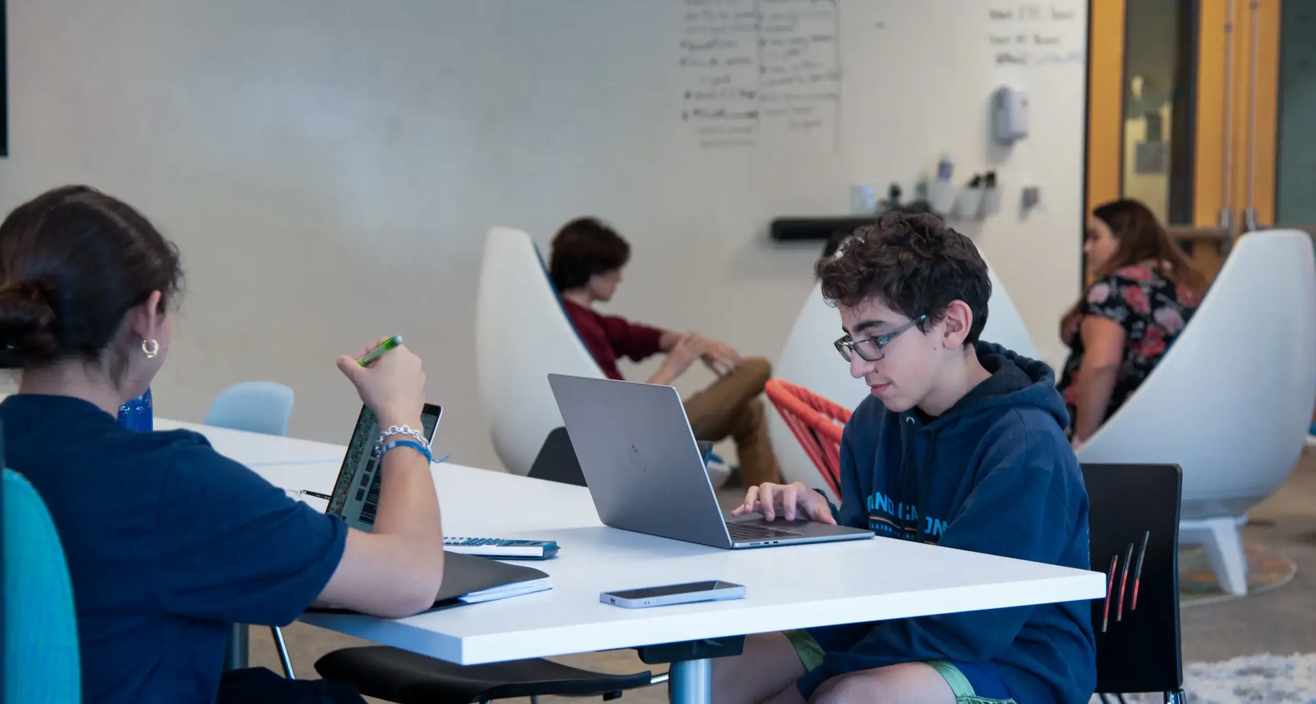 A group of students working in a modern, collaborative space. One student is sitting at a table using a laptop, while another is holding a green pen and looking at a notebook. In the background, two o