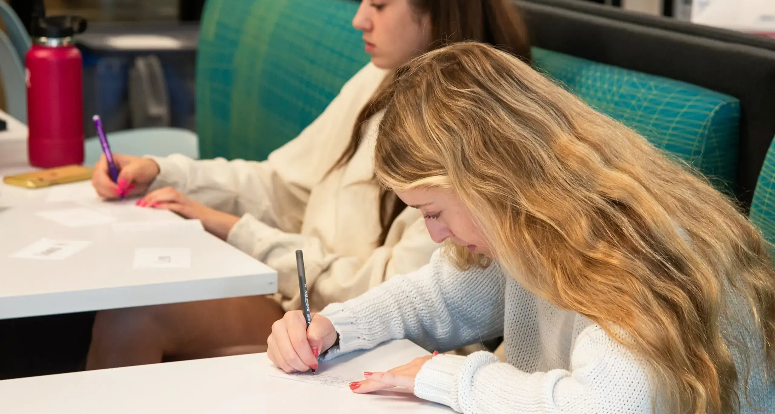 Two students are seated at a table in a classroom or study area, writing on pieces of paper. One student has long blonde hair and is focused on her writing, holding a purple pen. The other student, in