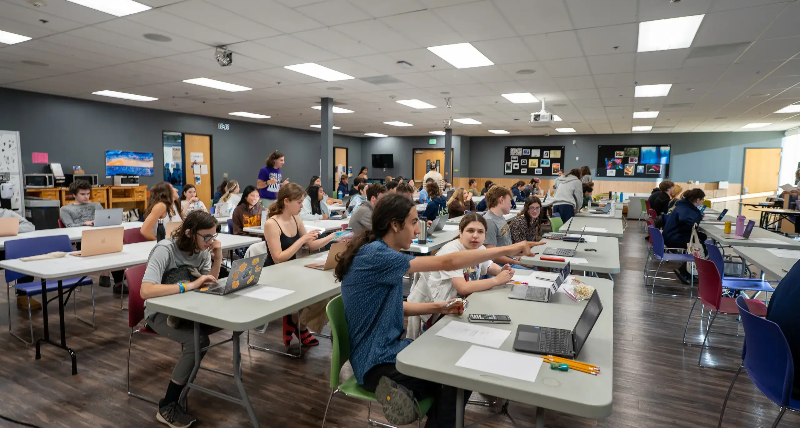 A large room with students seated at tables, working on laptops and interacting with each other. Walls display various art and educational materials.