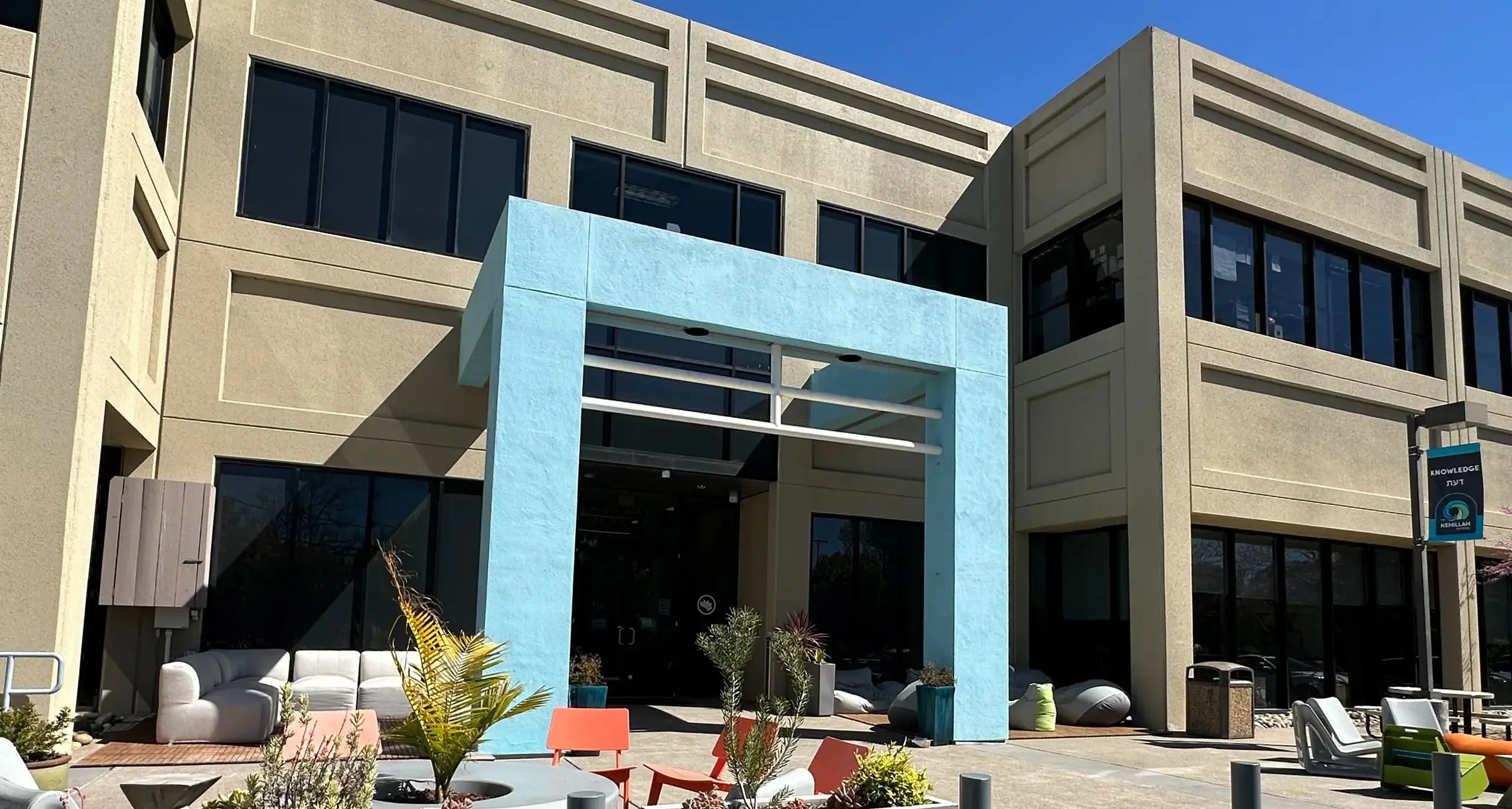 Exterior view of The Kehillah School – a modern building with large windows, a light blue archway at the entrance, and colorful outdoor furniture surrounded by planters on a sunny day.